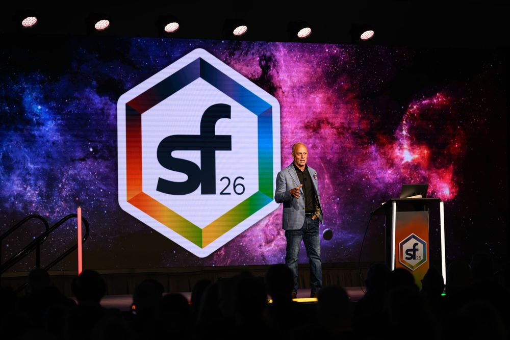 Speaker presents on stage at SF26 conference event with colorful hexagon logo backdrop.