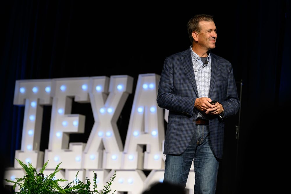 Man in blazer speaking on stage before illuminated TEXAS marquee letters at an indoor event.