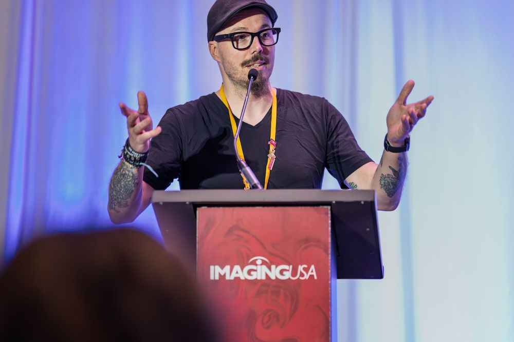Tattooed male speaker presenting at Imaging USA conference podium, gesturing with hands while wearing black shirt.