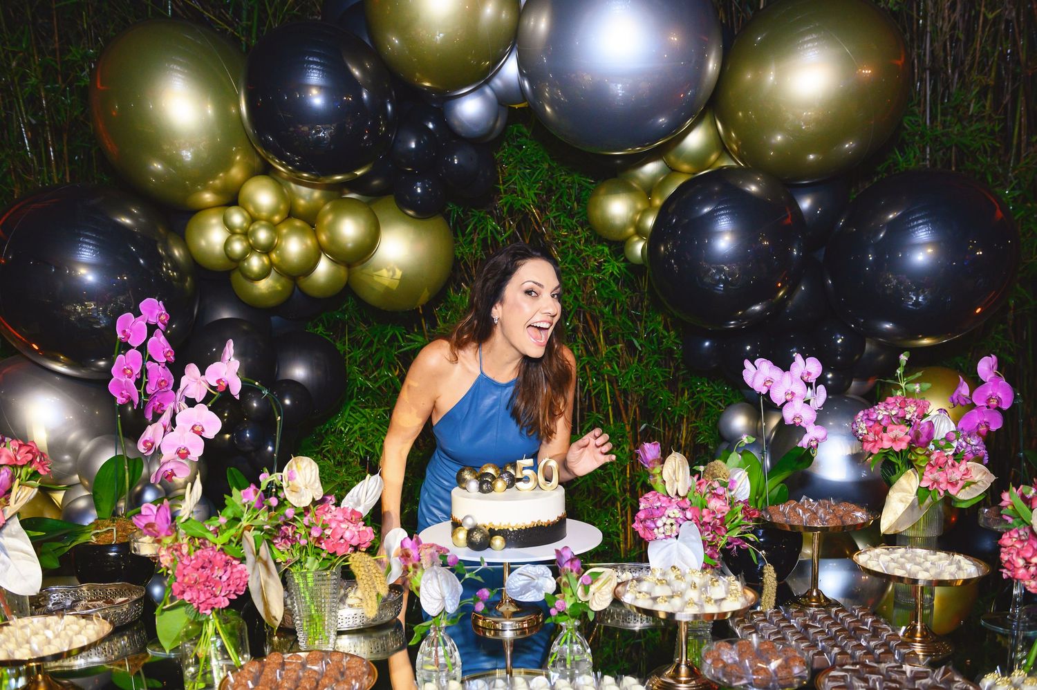 Woman celebrating birthday at elegant dessert table with black and gold balloons, pink orchids, and decorated cakes.