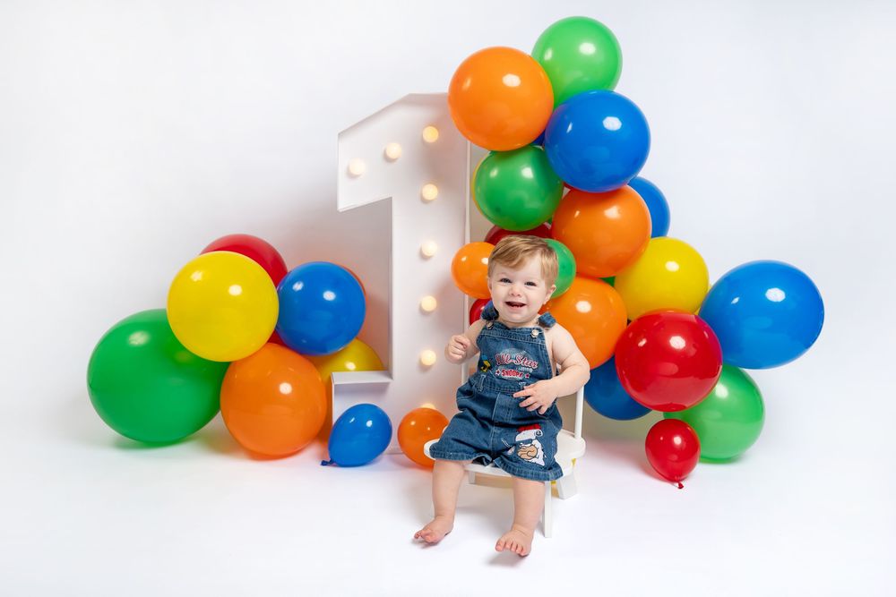 Baby girl in denim dress sits smiling before a large lit number 1 and colorful balloon arch on white background.
