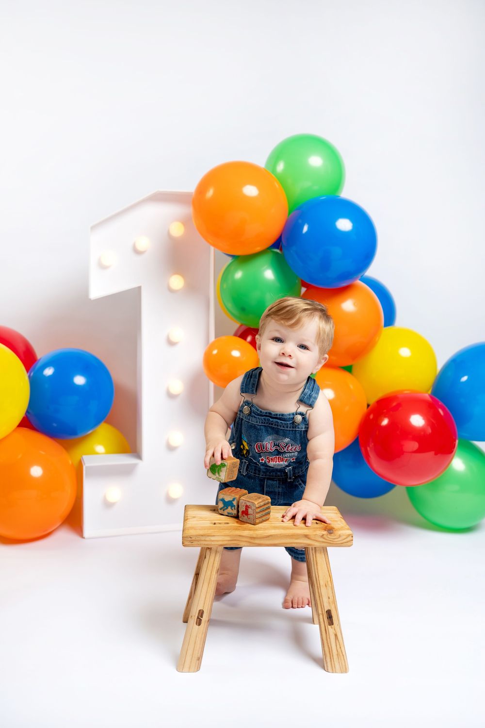 Happy toddler in denim overalls plays with wooden blocks on stool at colorful balloon first birthday setup.