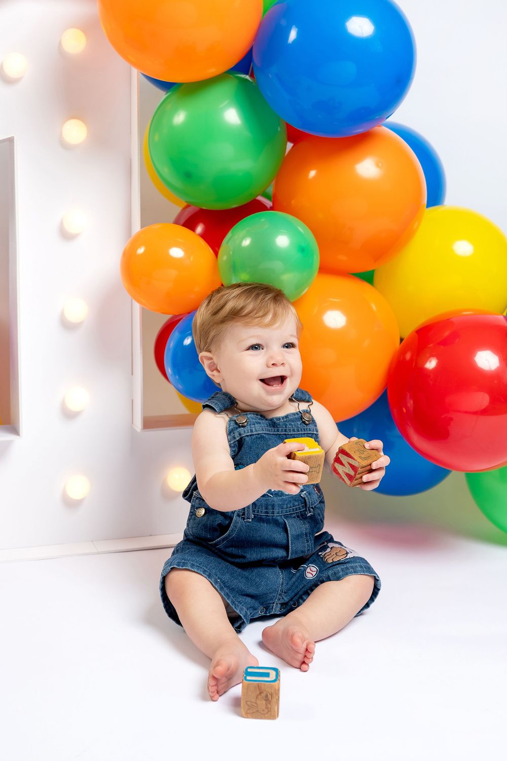 Happy toddler in denim overalls sitting on white floor with colorful balloons and illuminated number 1 backdrop.