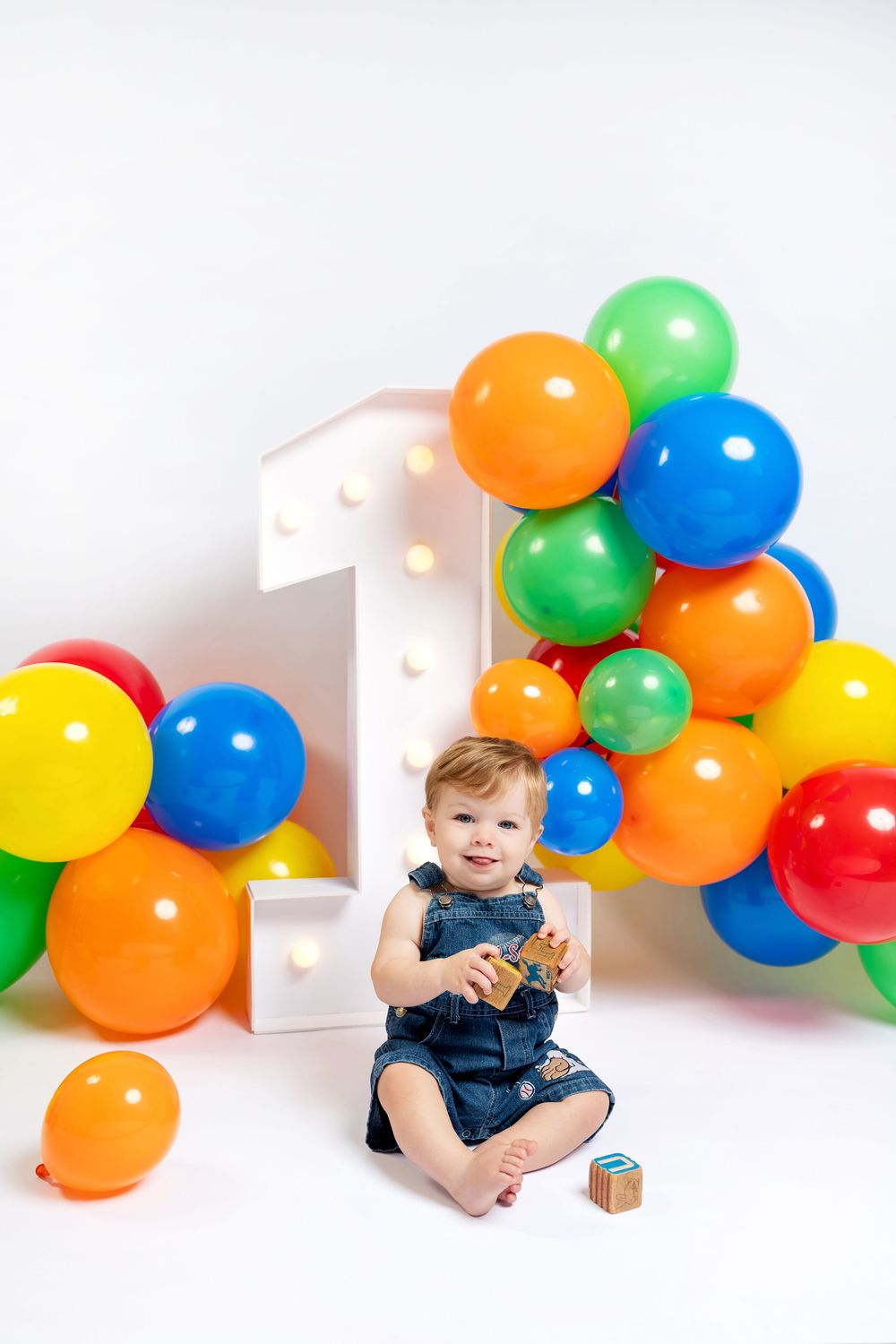 Baby girl in denim dress sits smiling at her first birthday party with colorful balloons and illuminated number one.