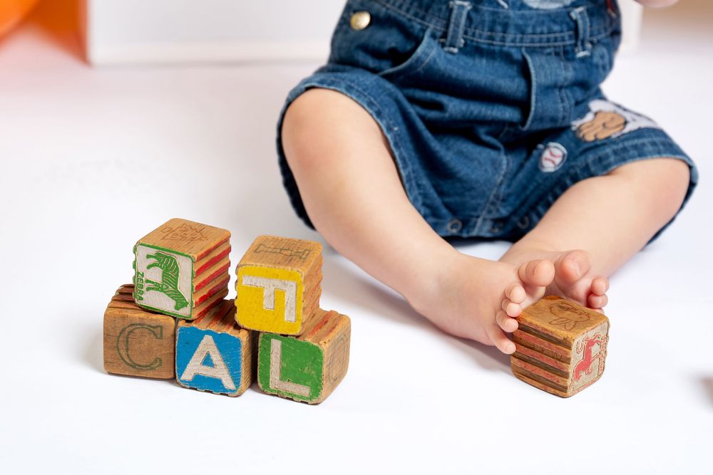 Baby in denim overalls playing with colorful wooden alphabet blocks on a white surface.