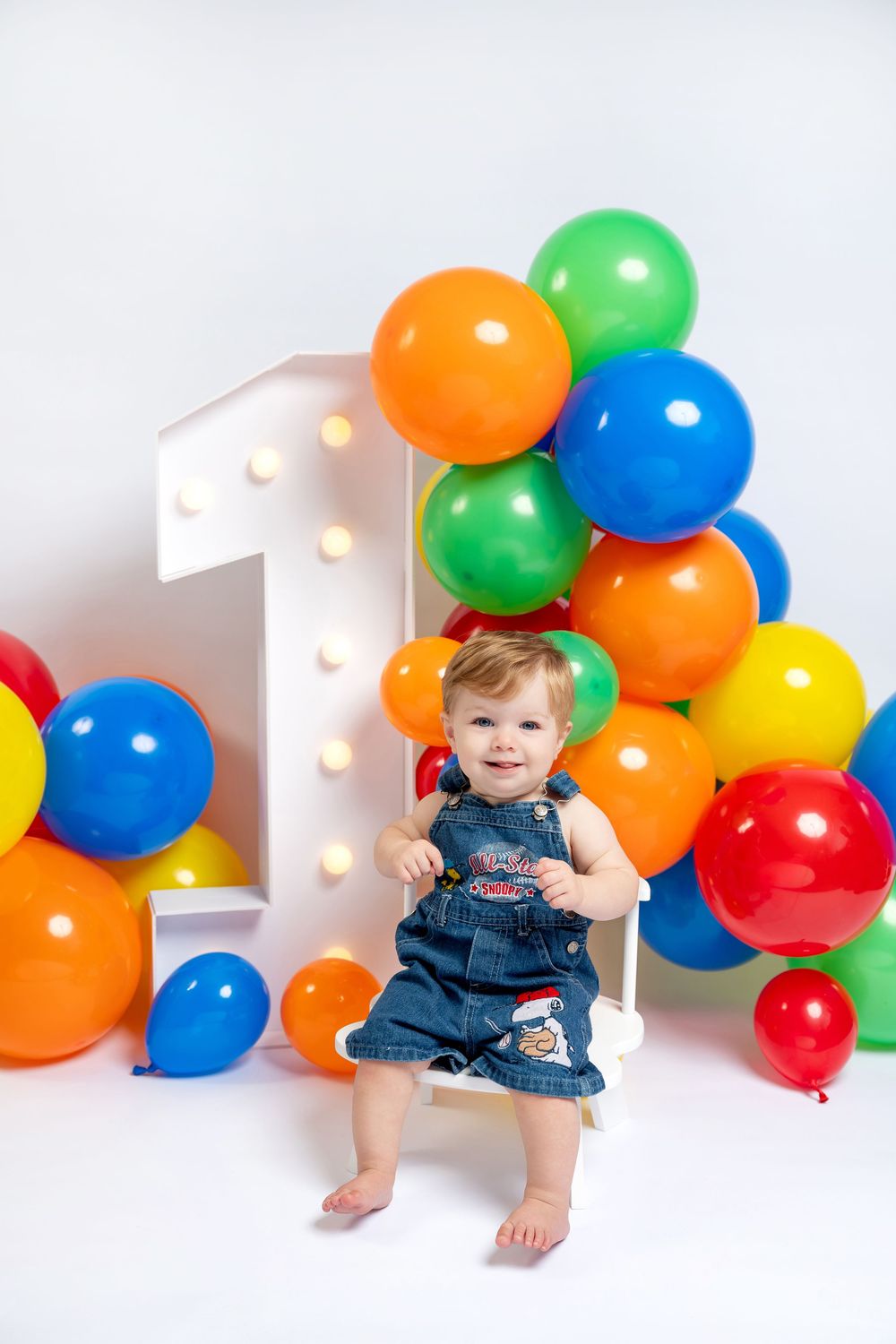 Smiling baby in denim overalls sits on white chair at 1st birthday party with colorful balloon arch backdrop.