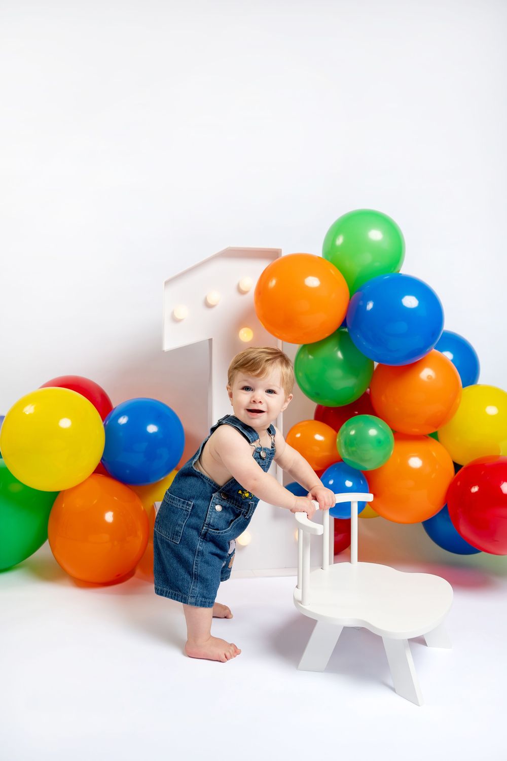 Smiling baby in denim overalls standing by a white number one with colorful balloons at a first birthday party.