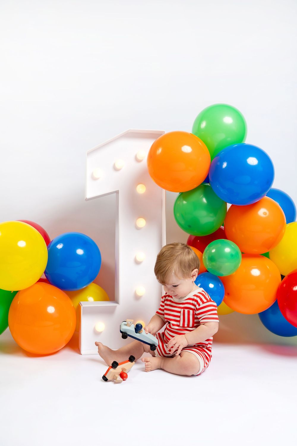 Baby's first birthday photo with colorful balloon arch, illuminated number one, and toy on white background.
