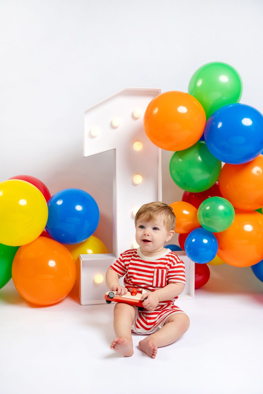 Baby in red striped outfit sits before colorful balloons and illuminated number 1, holding toy at first birthday celebration.