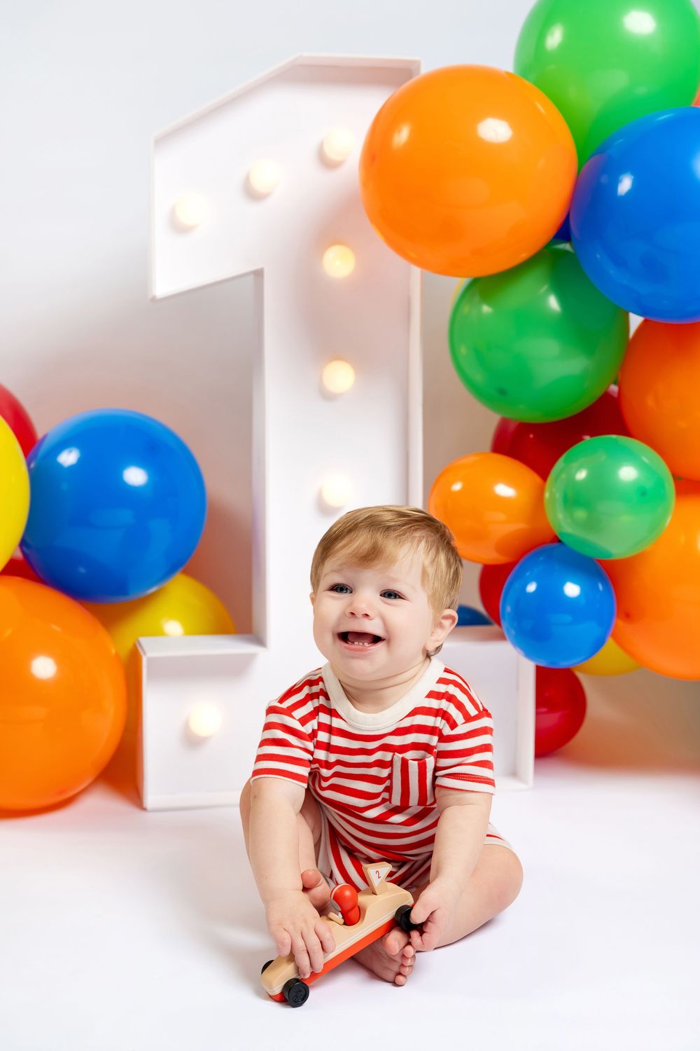 Smiling baby in red striped outfit celebrates first birthday with colorful balloons and illuminated number one decoration.