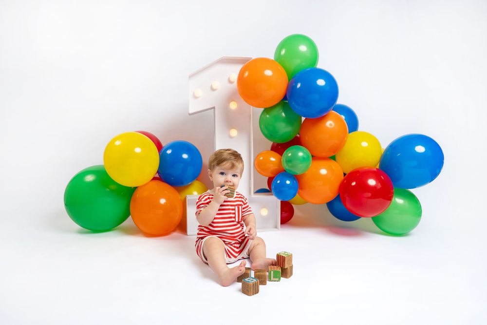 Baby in red outfit sits on floor with wooden blocks, colorful balloons and lit number 1 on white background.