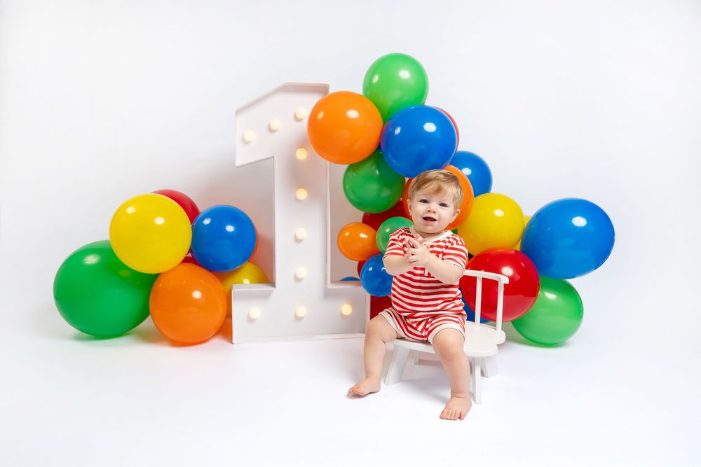 Happy baby in red striped outfit sitting on white bench with colorful balloon arch and number 1 backdrop.
