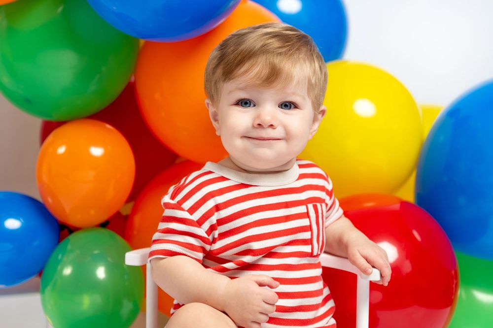 Smiling toddler in red striped shirt sitting among colorful birthday balloons on white background.