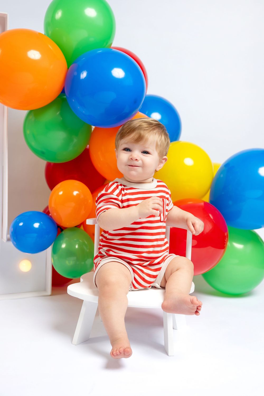 Smiling baby in red striped outfit sitting on white chair surrounded by colorful balloons at first birthday party.