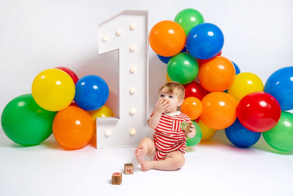 One-year-old baby in red striped romper playing with blocks beside colorful balloon arch and number 1 decor.