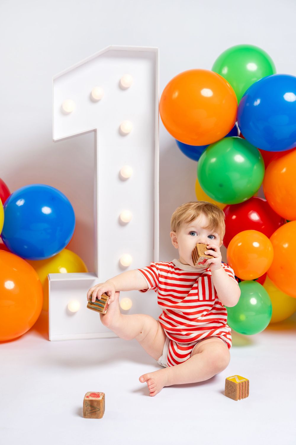Baby in red striped outfit celebrating 1st birthday with colorful balloons and wooden blocks.