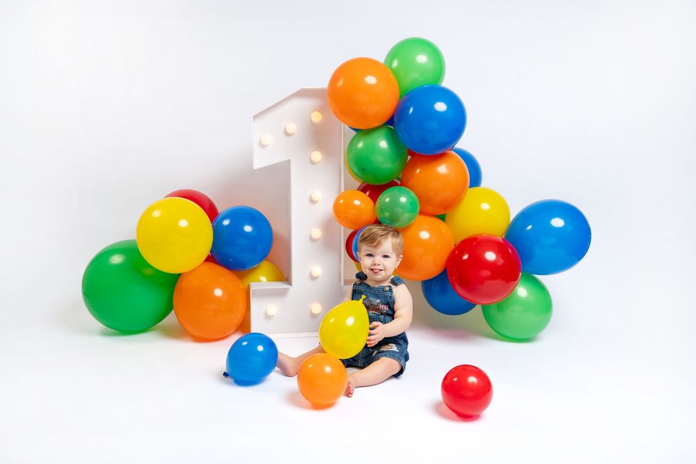 Baby's first birthday photo with colorful balloons and a large number one on a white background.