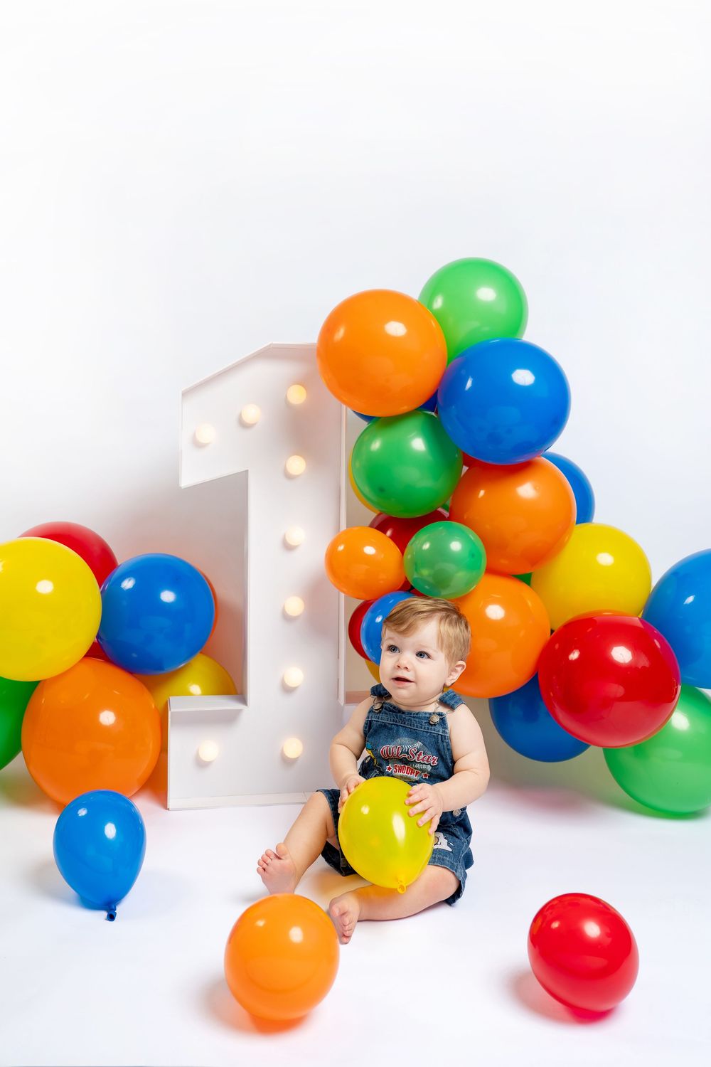 Baby sitting on white floor surrounded by colorful balloons celebrating first birthday with large number one decoration.