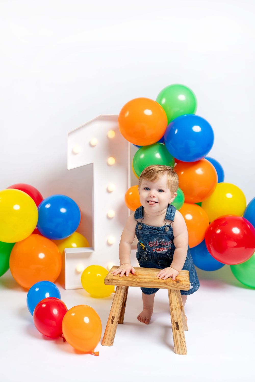 Smiling toddler in denim overalls stands by wooden stool with colorful balloons and illuminated number 1 on white background.