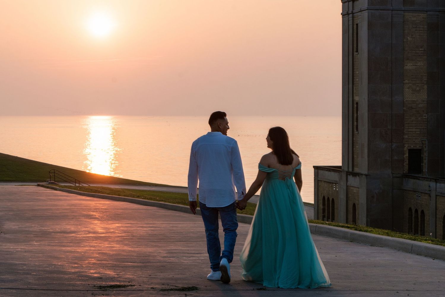 Couple holding hands watching a romantic sunset over the ocean, woman in teal gown, man in white shirt.