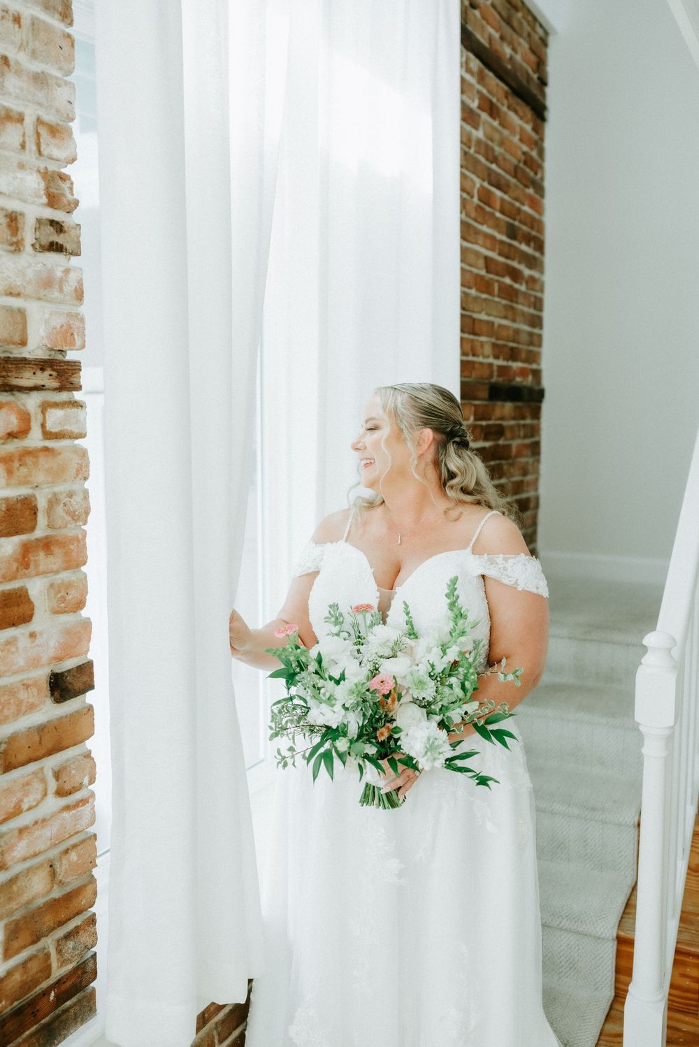 Bride in off-shoulder white gown holds floral bouquet by sheer curtains near exposed brick wall.