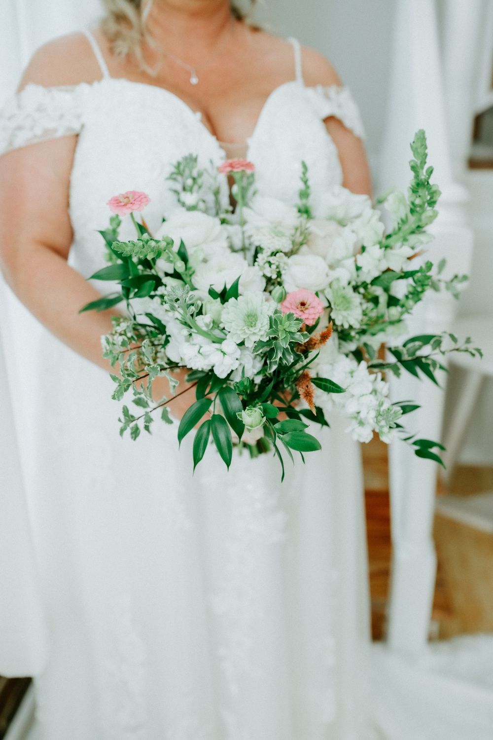 Bride in lace off-shoulder gown holding lush white and pink floral bouquet with greenery.