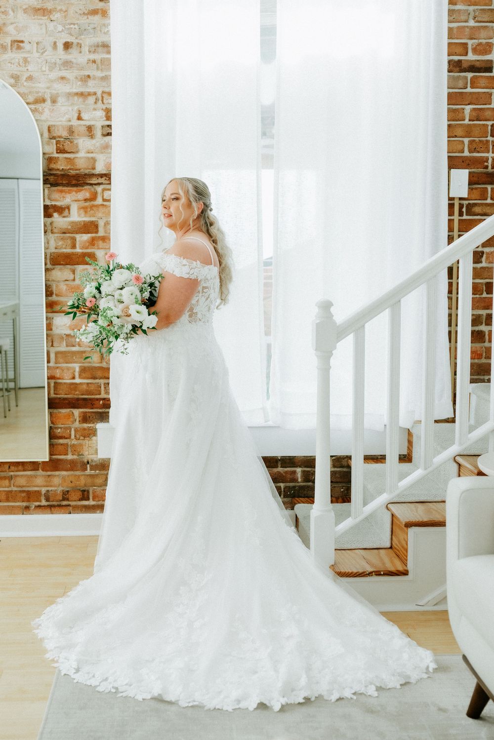 Bride in off-shoulder lace wedding gown with train holds bouquet near brick wall and white staircase.