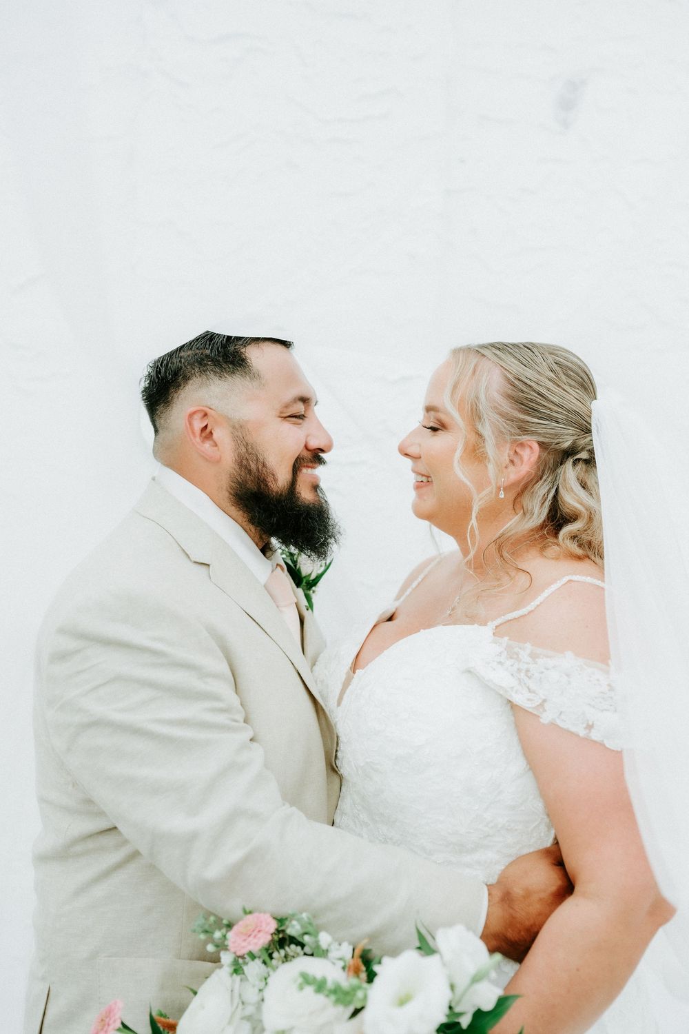 A bride and groom share a joyful moment together, smiling at each other, dressed in wedding attire against a white wall.