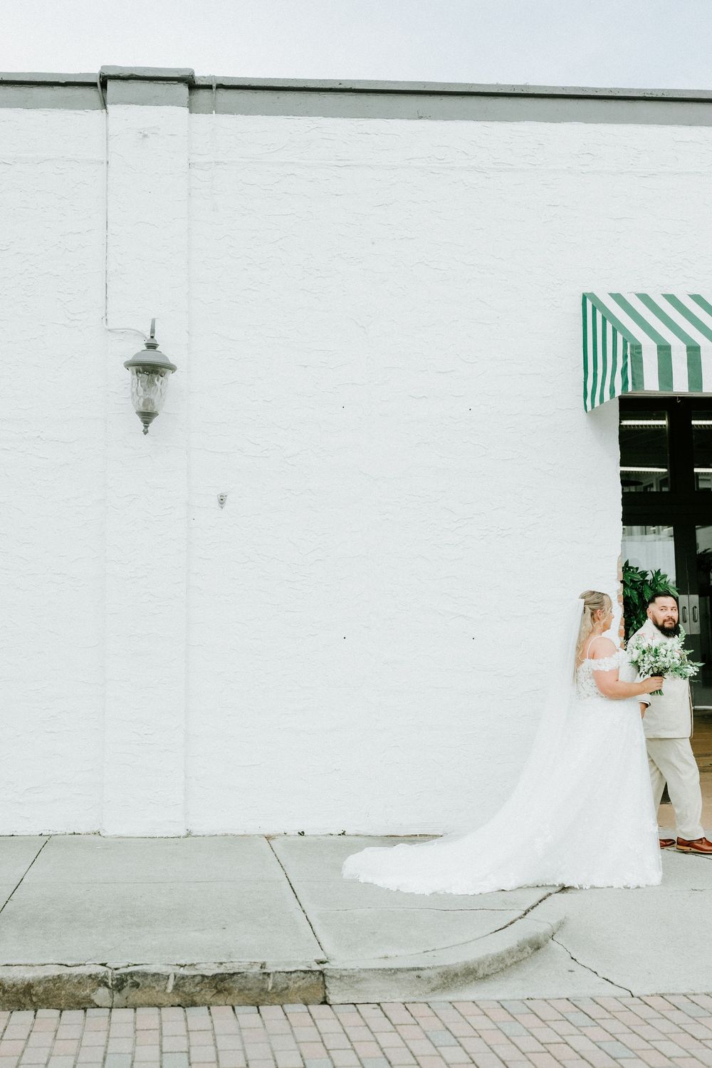 Bride and groom share a romantic moment outside a white brick building with a green striped awning on their wedding day.