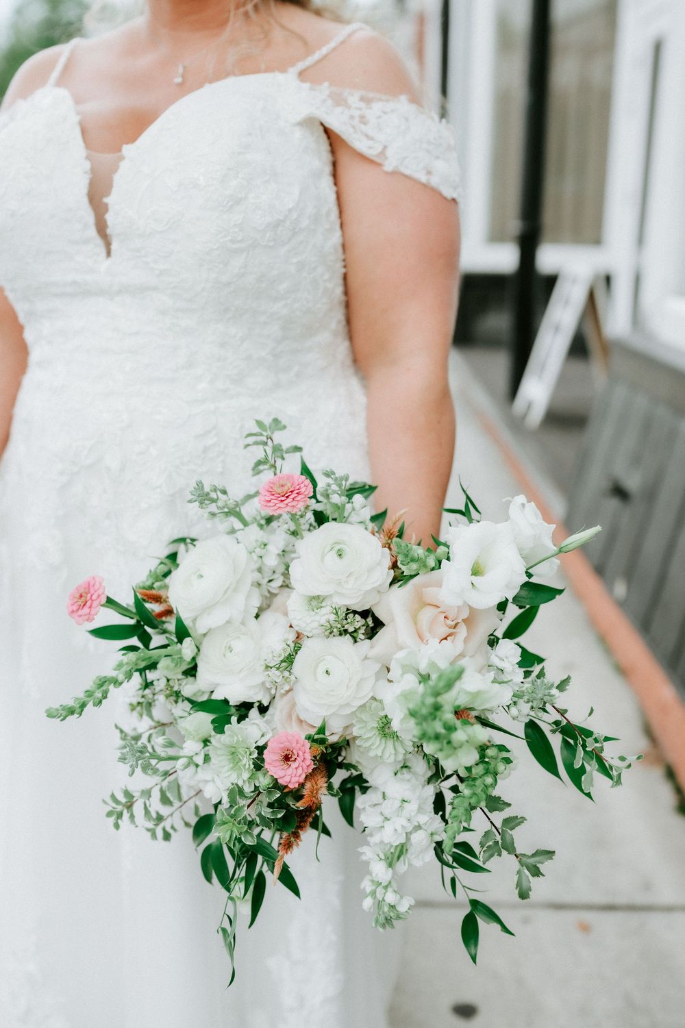 Bride in lace off-shoulder wedding gown holding lush bridal bouquet with white ranunculus, pink flowers, and greenery.