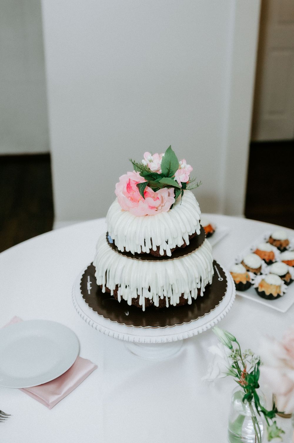 Two-tier white bundt wedding cake with pink floral topper on white cake stand, surrounded by mini desserts on draped table.