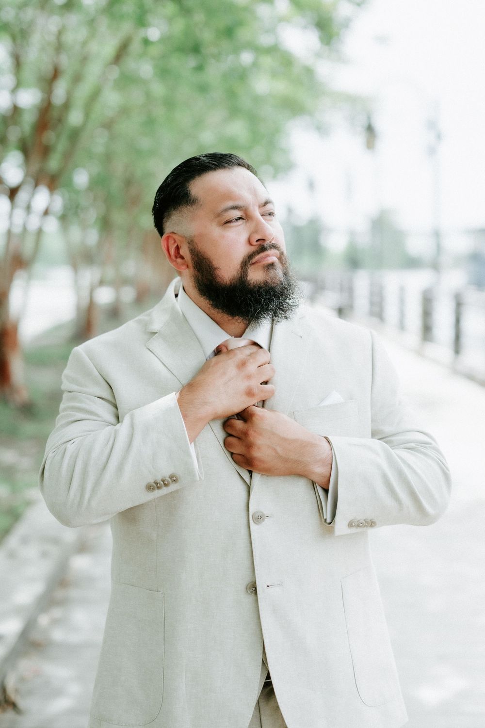 A bearded man in a light gray suit adjusts his tie outdoors near trees and a waterway.