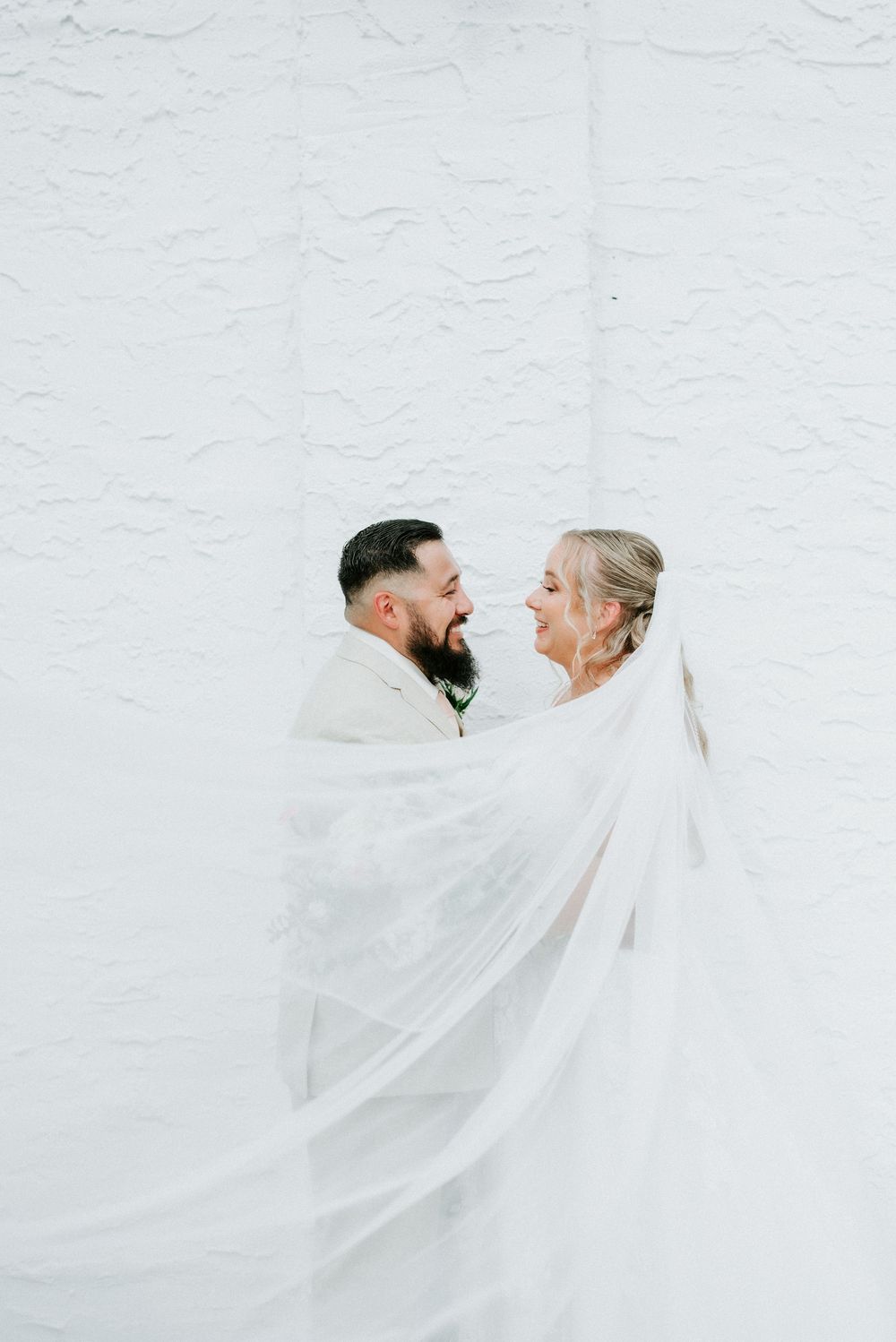 Bride and groom wrapped in a flowing veil, sharing a joyful moment against a white textured wall.