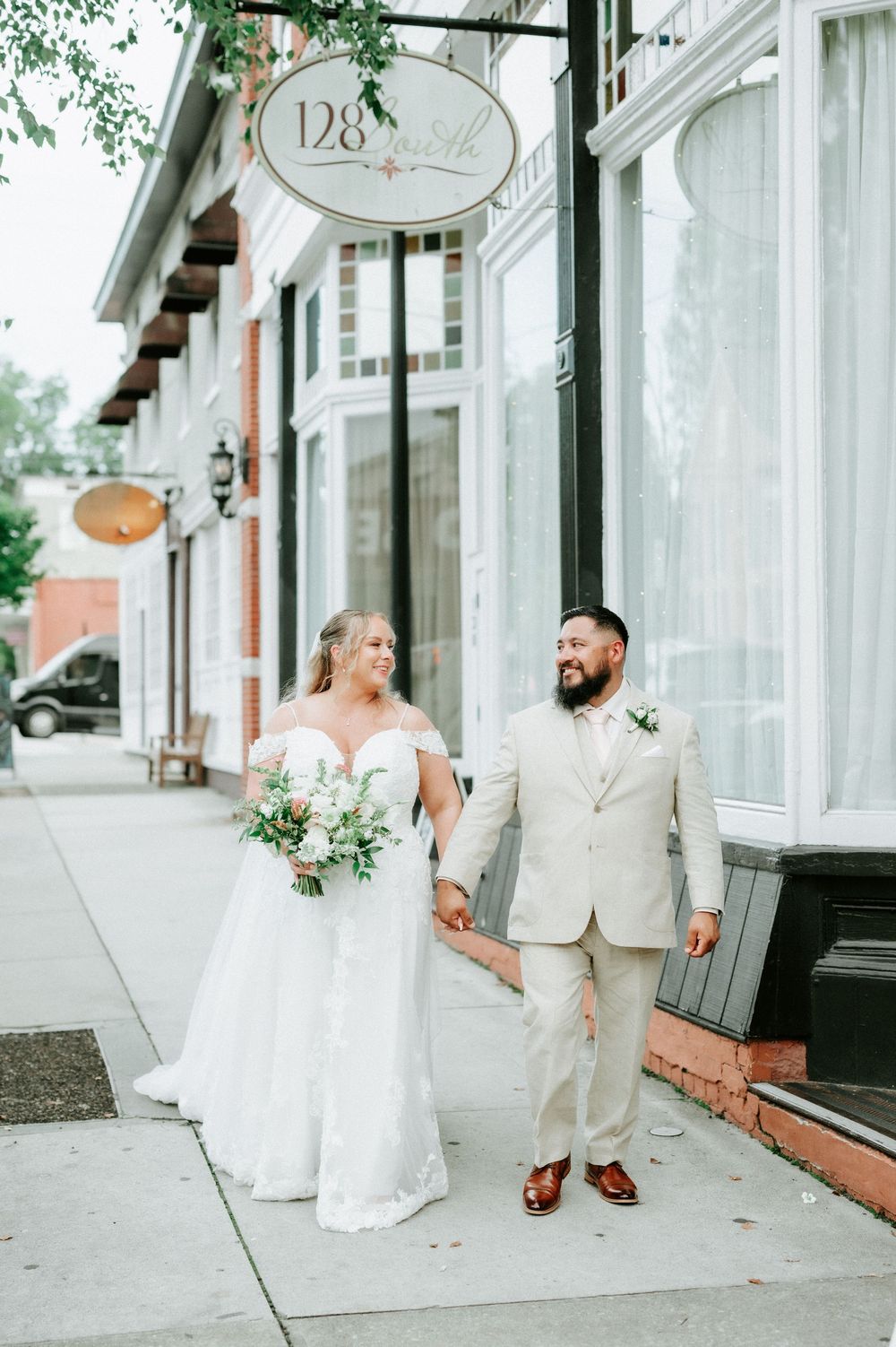 Bride and groom walking hand in hand on a sidewalk outside 128 South venue, bride holding a bouquet.