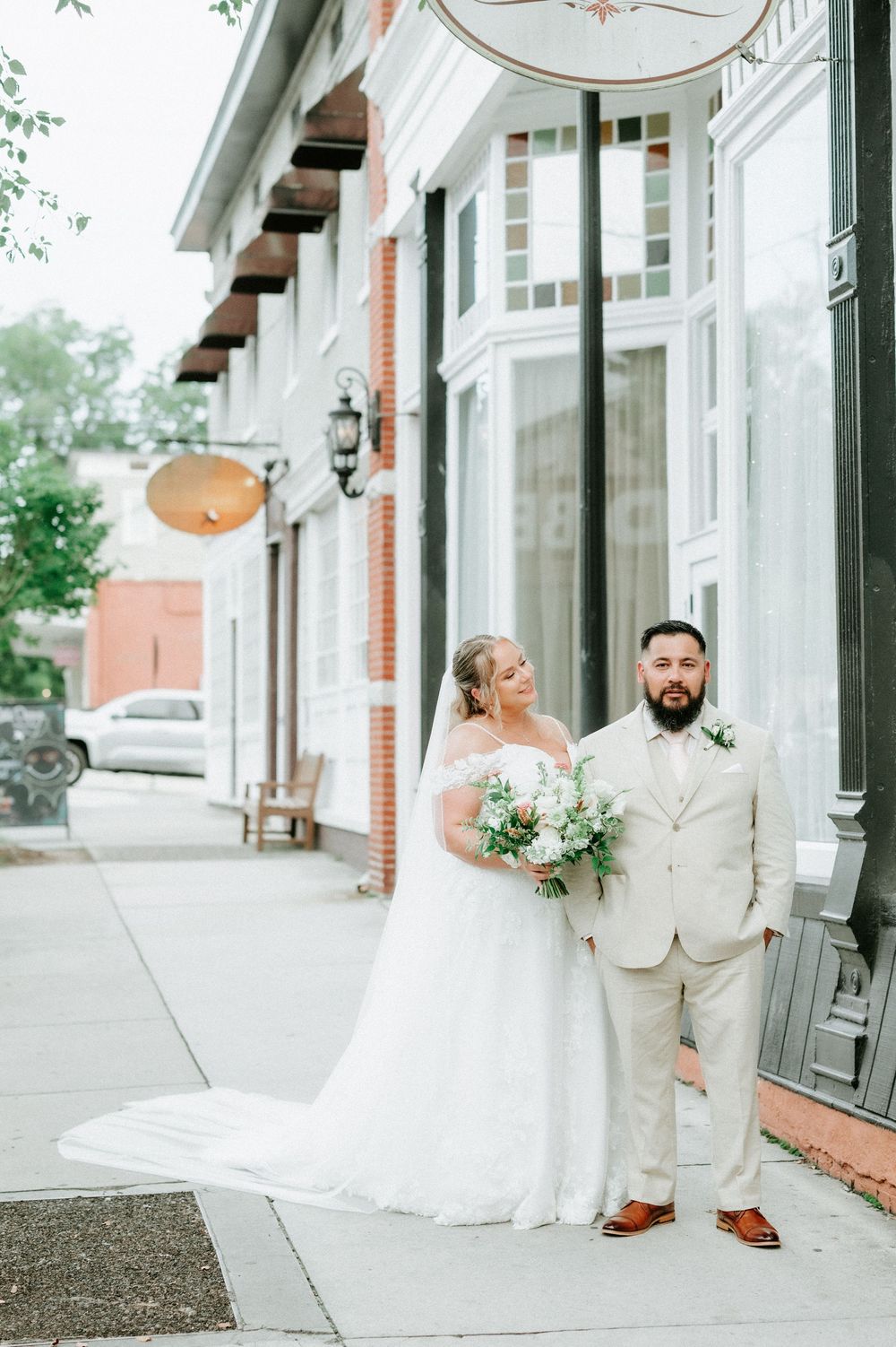 Bride in white gown holding bouquet stands with groom in tan suit on sidewalk outside brick building.