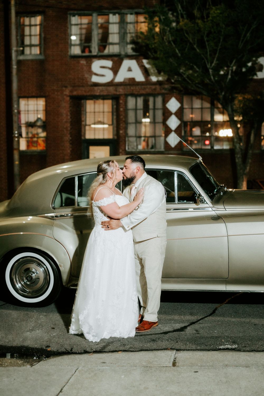 Bride and groom share a romantic kiss beside a vintage gold Rolls-Royce at night outside a brick building.