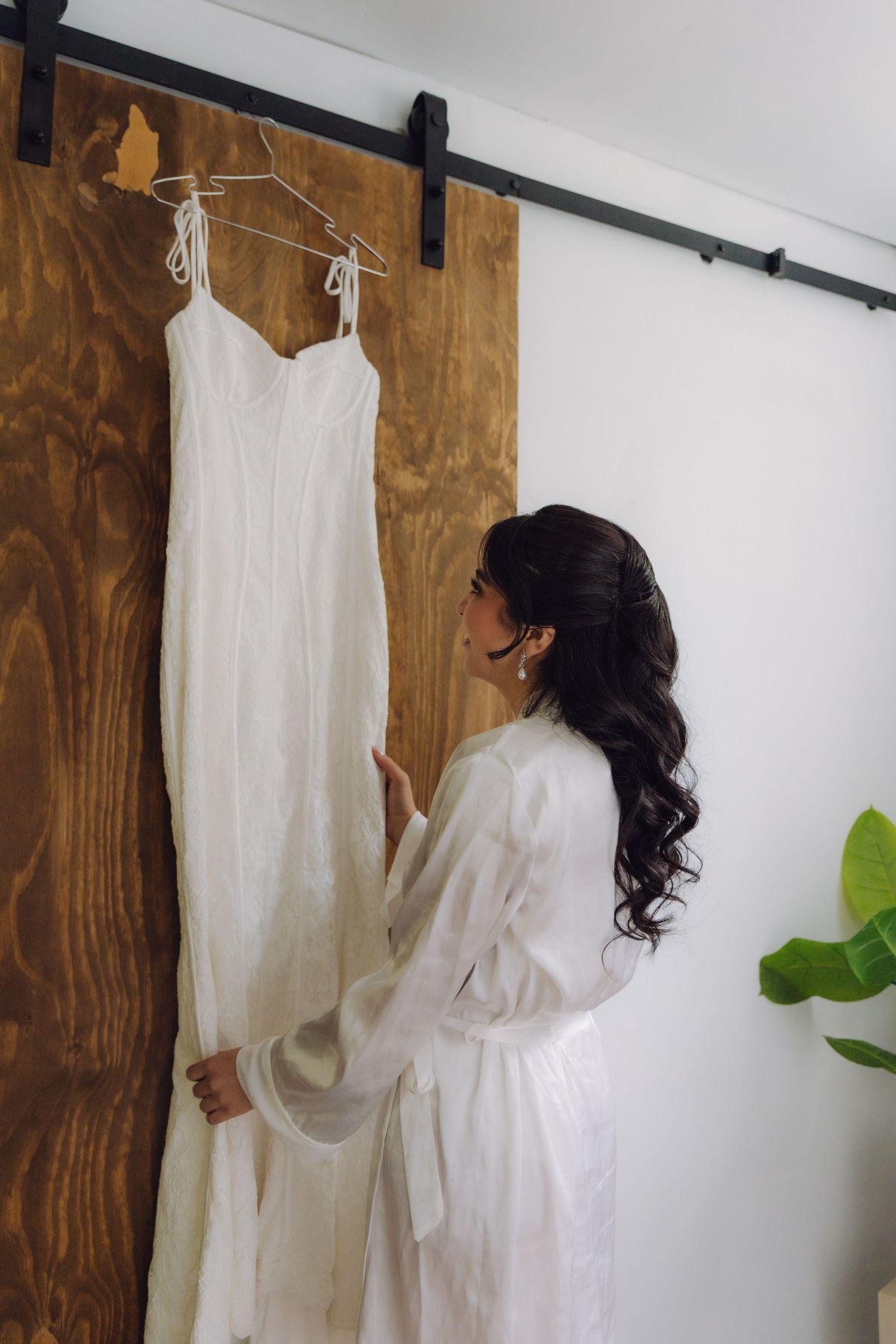 Bride in white robe admires her wedding dress hanging on a rustic barn door before getting ready.