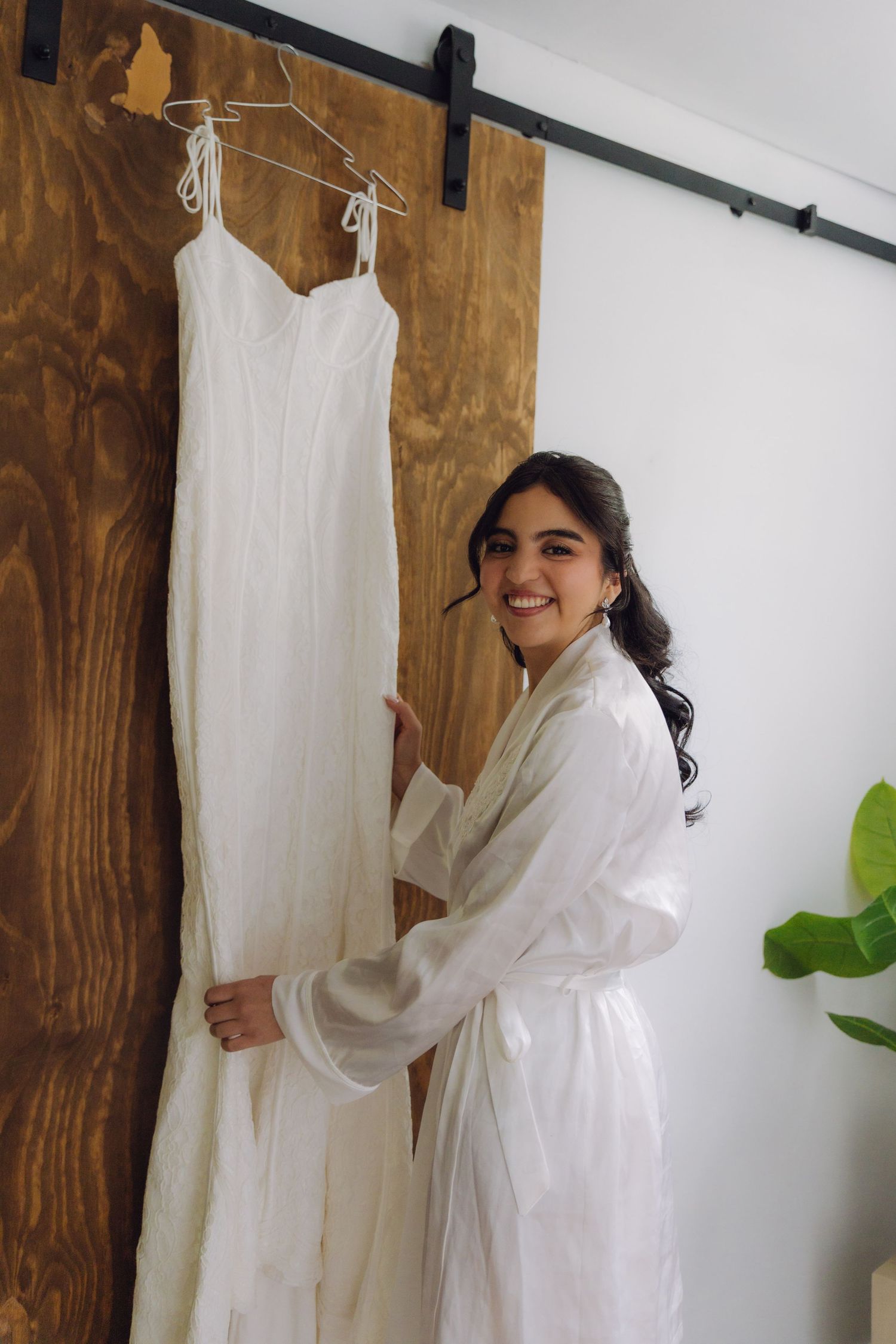 Smiling bride in white robe stands beside her elegant wedding gown hanging on a barn door.