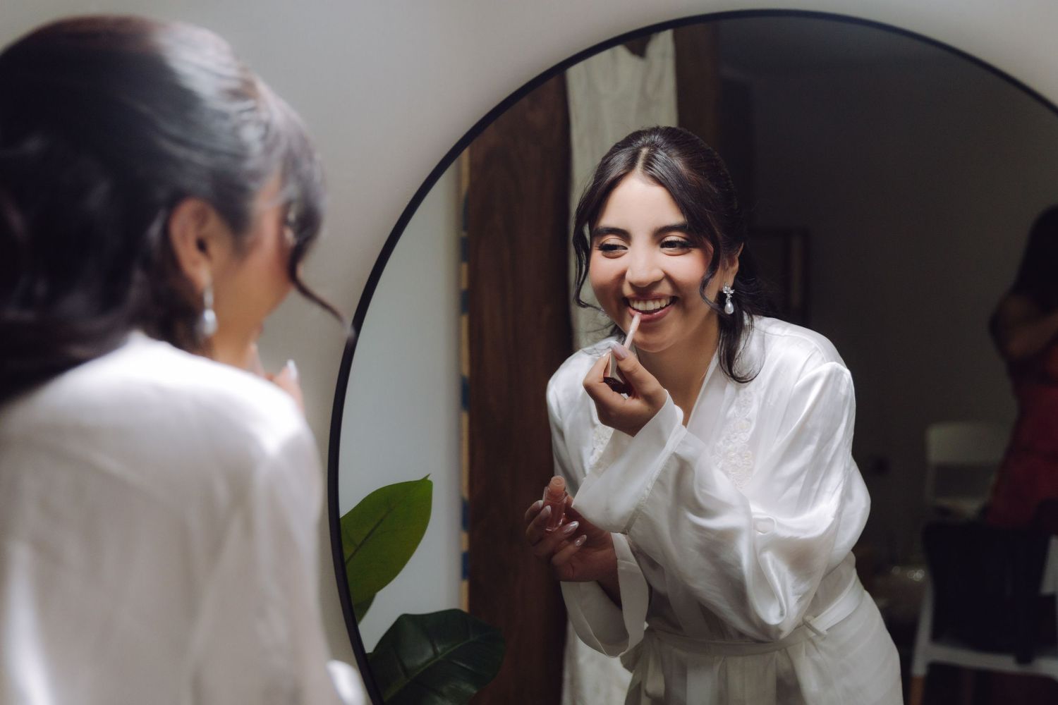 Happy bride in white robe applies lipstick while smiling at her reflection in a round mirror.