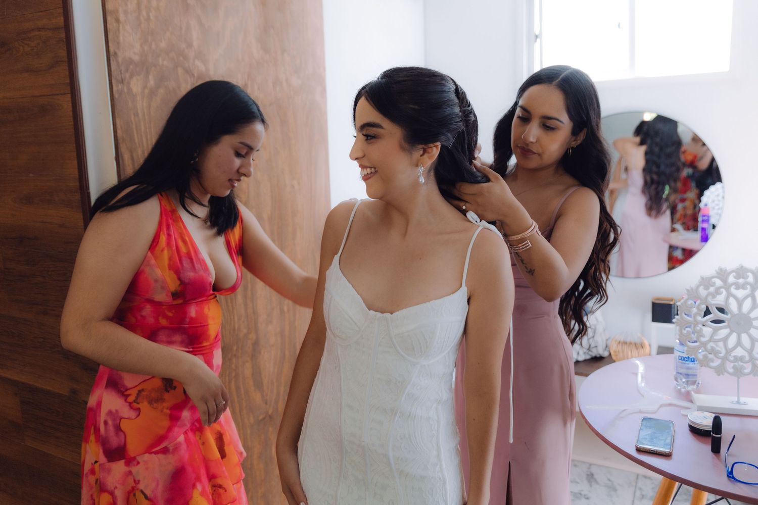 Two bridesmaids help the smiling bride with final touches to her hair and wedding dress.
