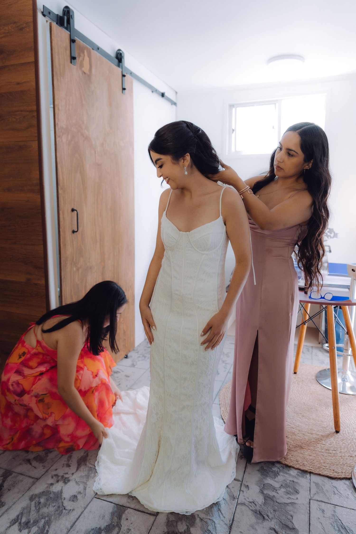 Two bridesmaids assist the bride in her fitted white lace gown near a rustic barn door.