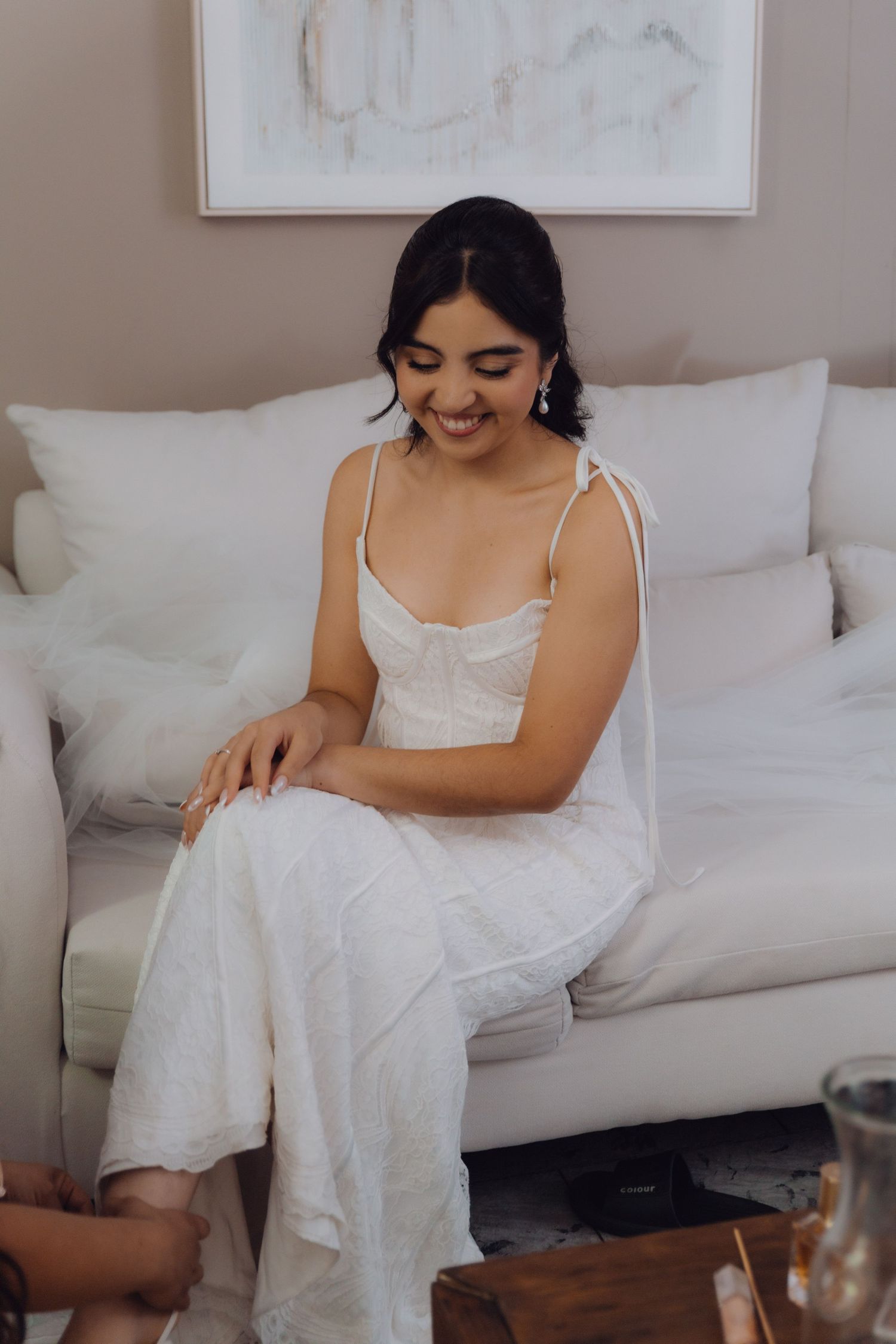 Radiant bride in a white lace gown sits on a white sofa smiling during wedding preparations.