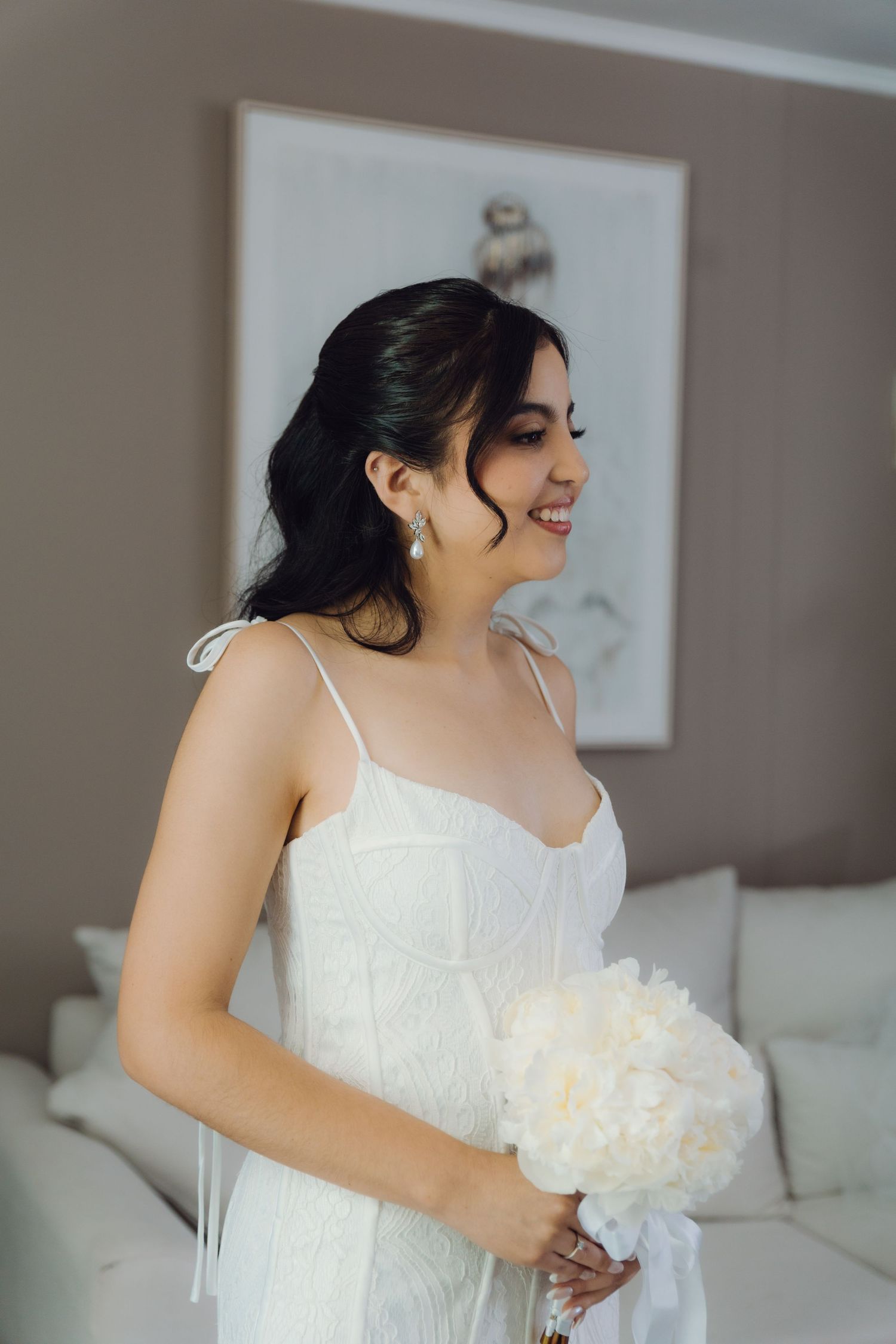 Smiling bride in white spaghetti strap wedding gown holding white peony bouquet indoors.