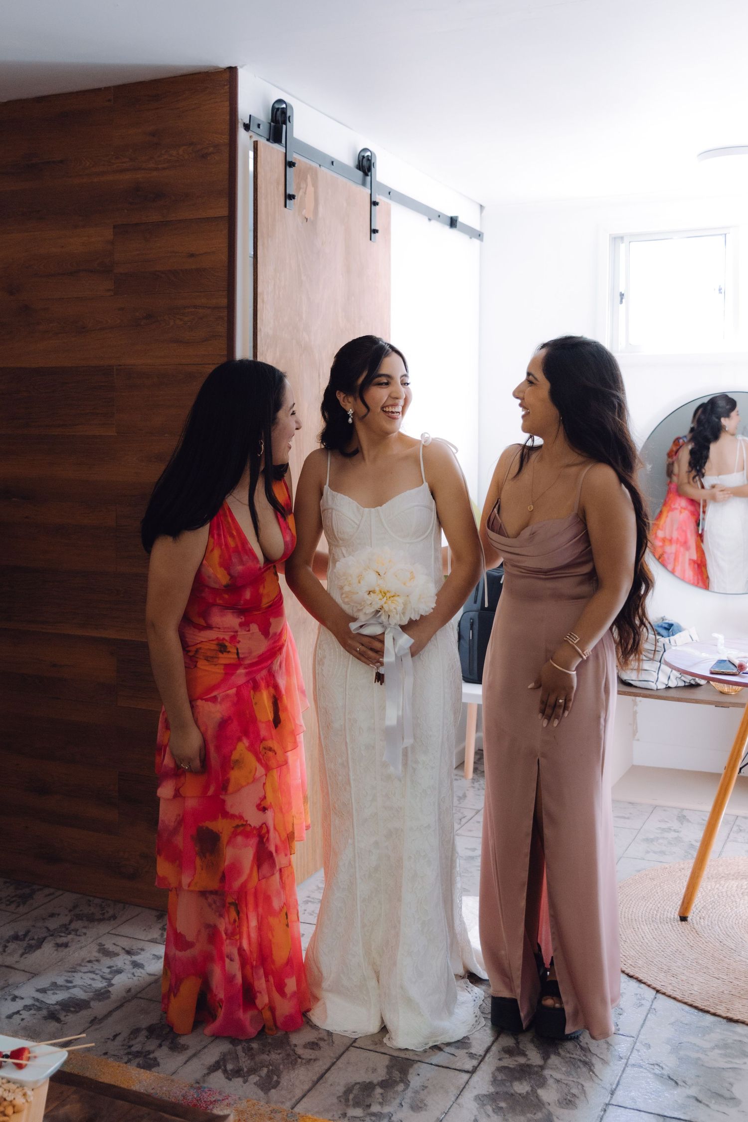 Bride in white gown laughing with two bridesmaids in colorful dresses near barn door.
