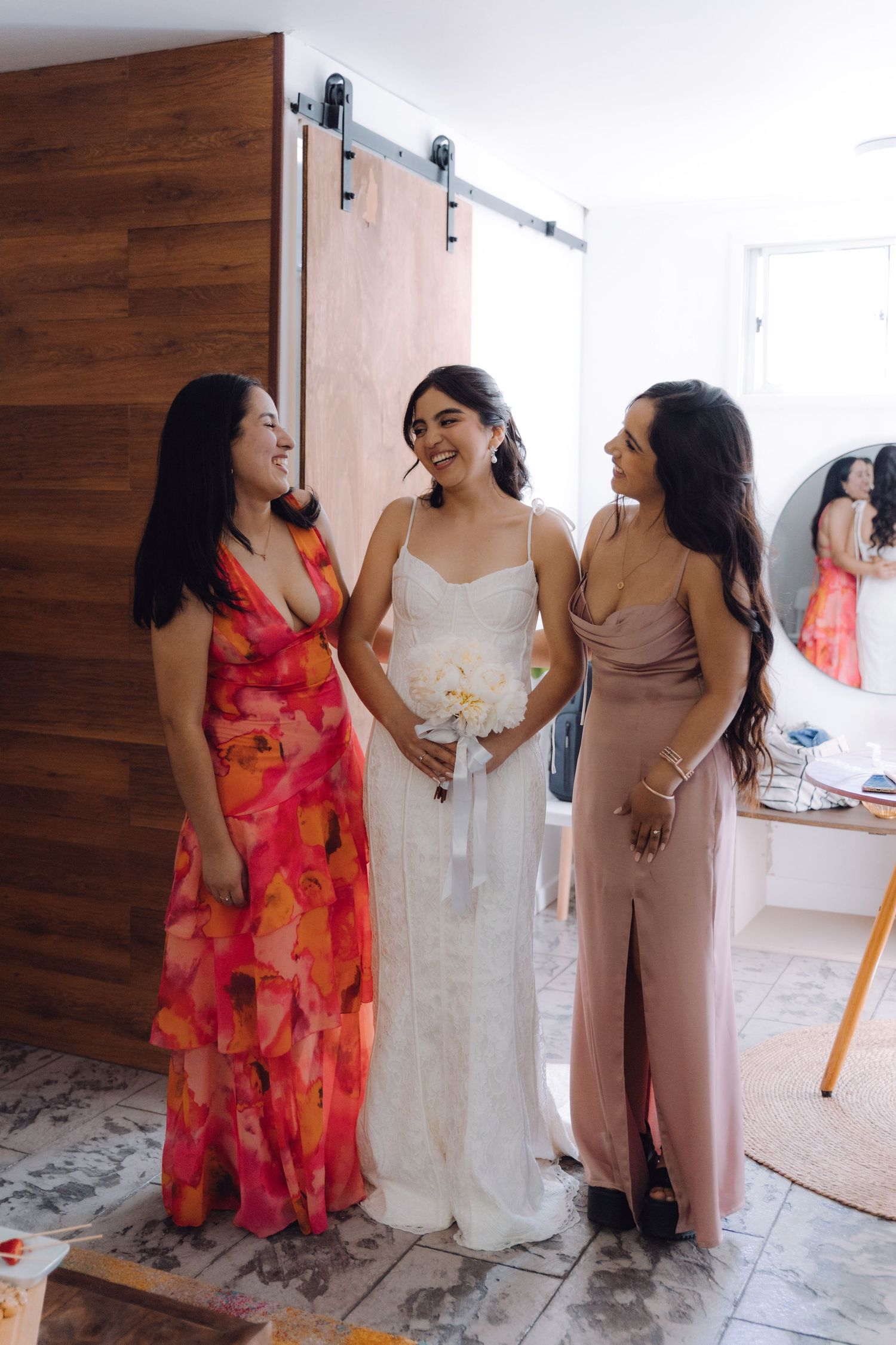 Bride holding white bouquet smiling with bridesmaid in pink and bridesmaid in floral dress.