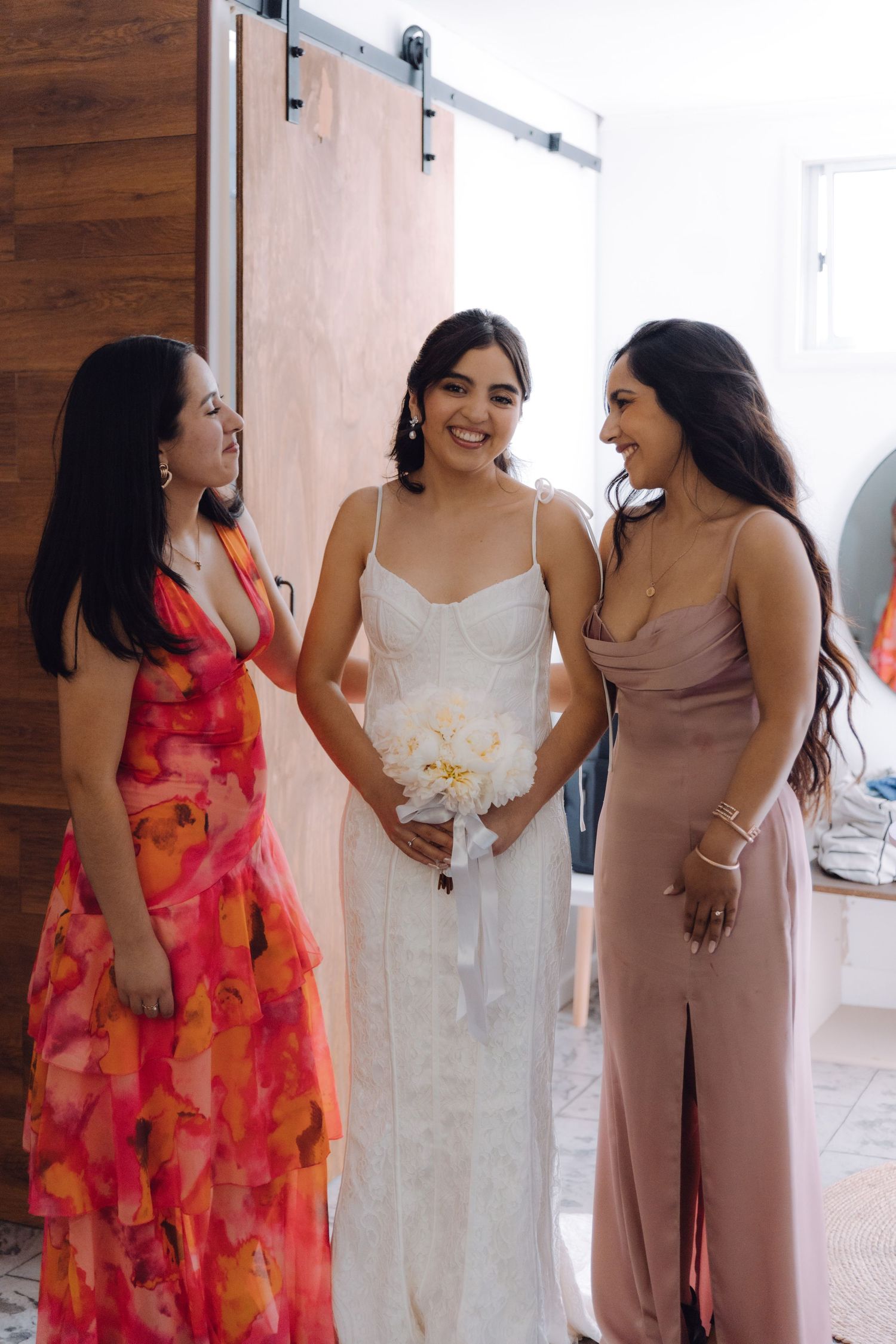 Joyful bride in fitted white dress laughing with two bridesmaids in elegant gowns indoors.
