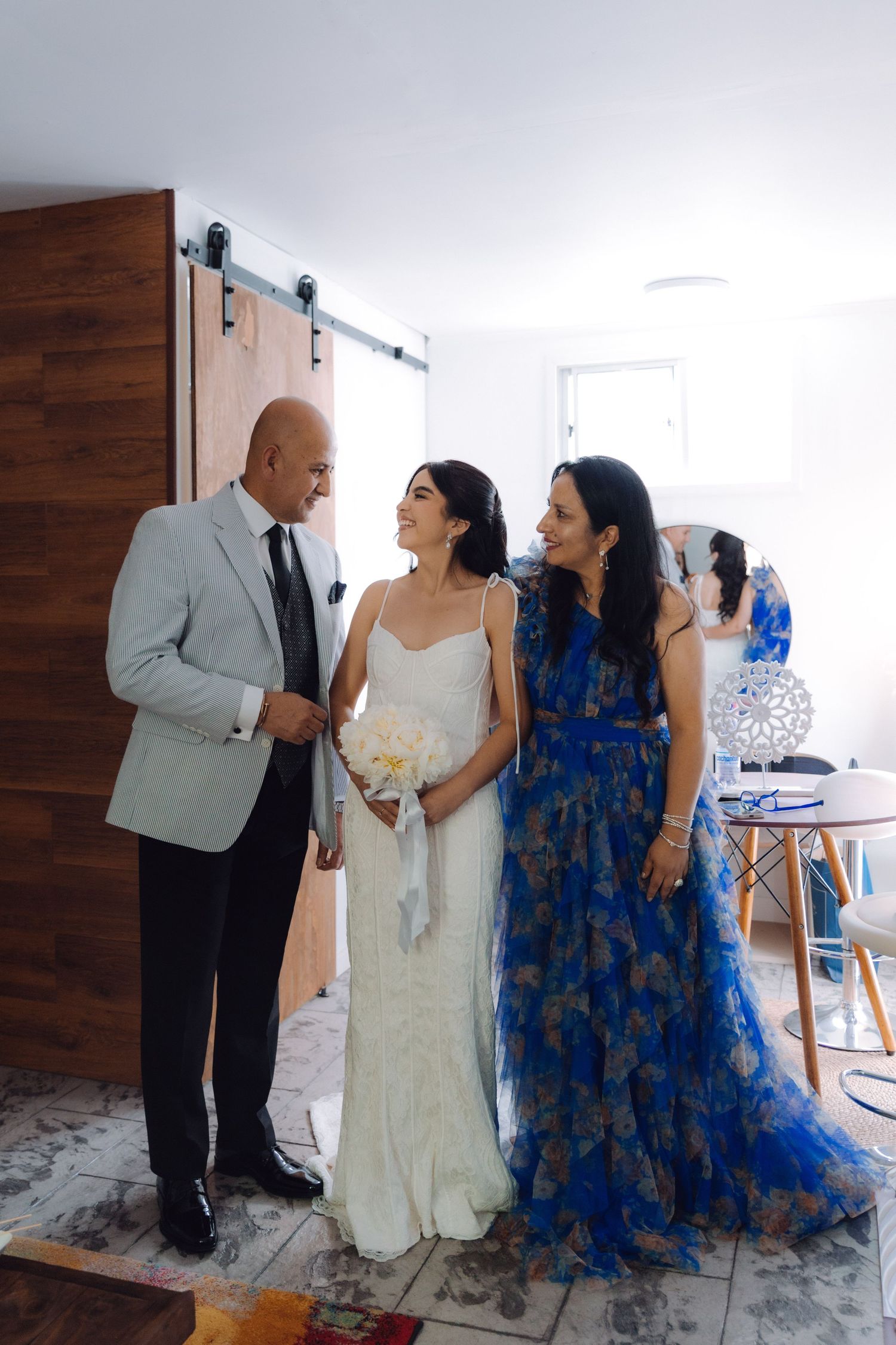 Bride in white gown smiling with parents, father in suit and mother in blue floral dress.