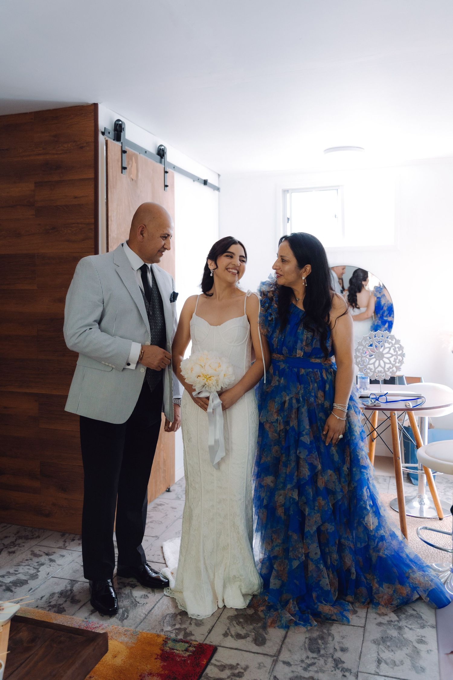 Bride laughing with parents near barn door, mother wearing vibrant blue floral gown.