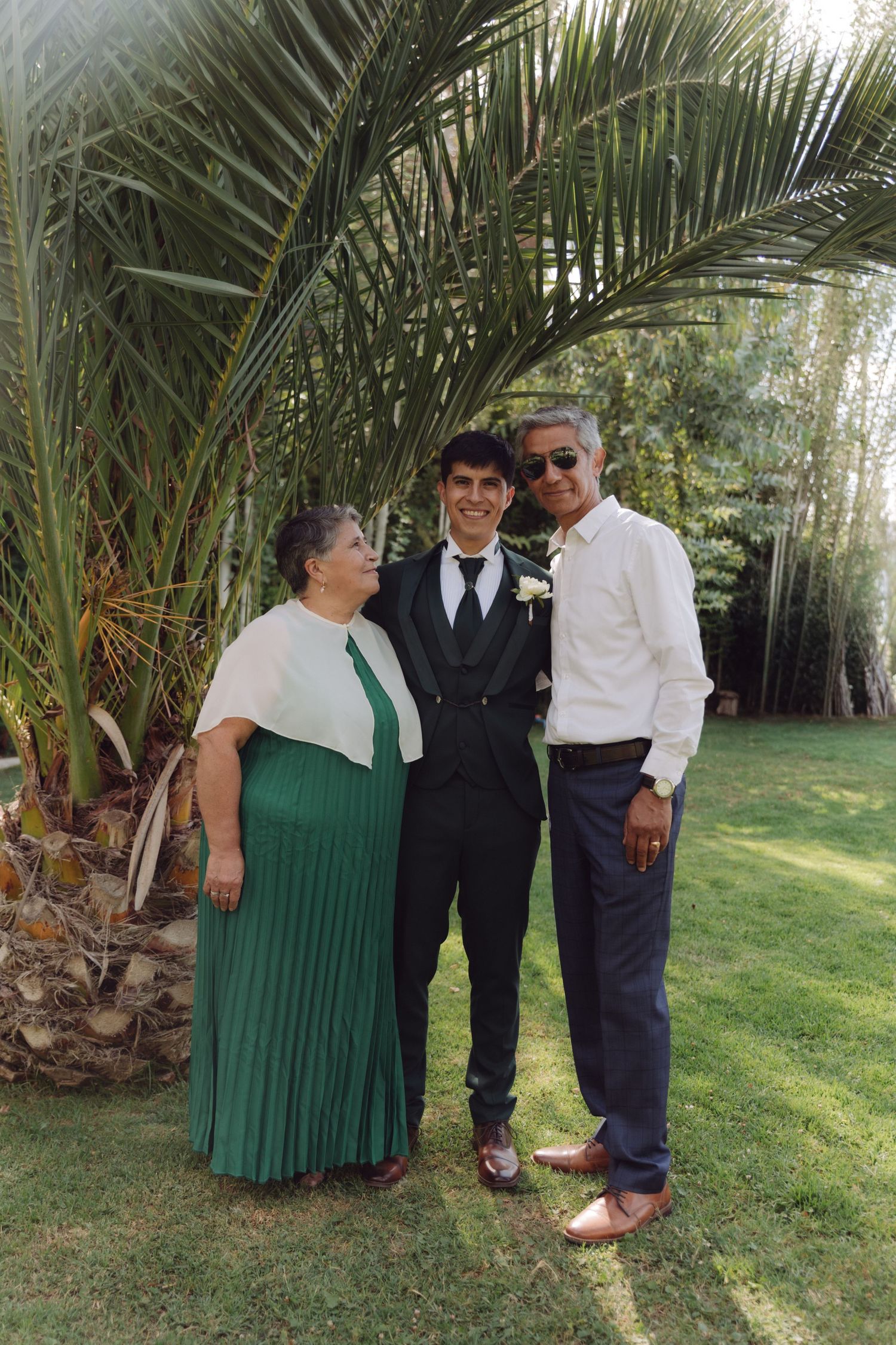 Groom in black suit posing with parents under palm tree at outdoor wedding venue.