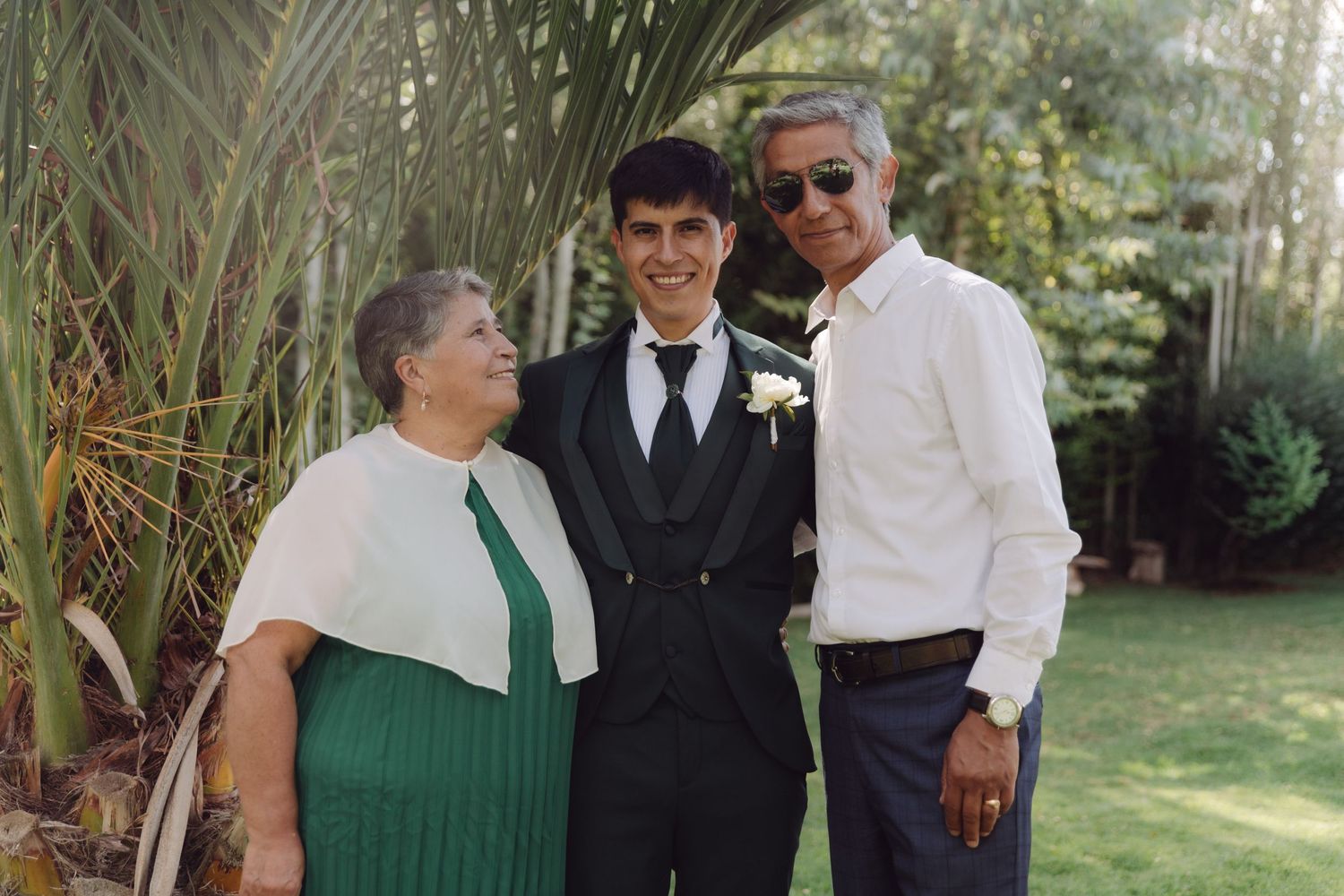 Groom in black tuxedo smiling with parents outdoors at garden wedding celebration.